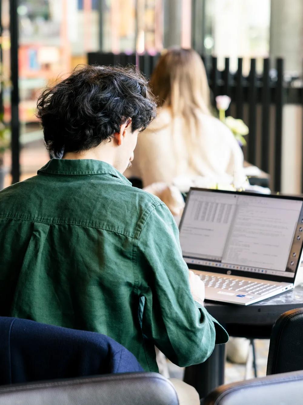 A person with dark hair wearing a green shirt works on a laptop at a round table in a bright, modern indoor space. Other people are visible in the background.