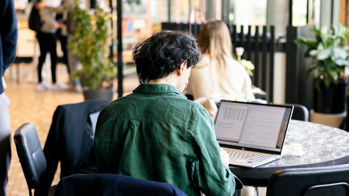 A person with dark hair wearing a green shirt works on a laptop at a round table in a bright, modern indoor space. Other people are visible in the background.