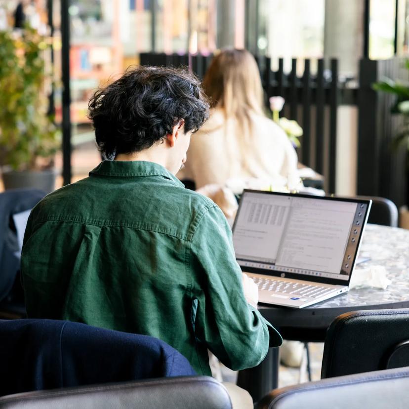 A person with dark hair wearing a green shirt works on a laptop at a round table in a bright, modern indoor space. Other people are visible in the background.