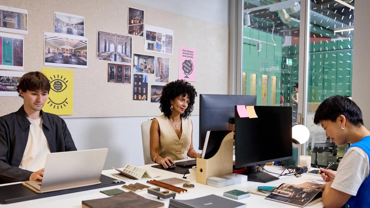 Drei Personen arbeiten an einem gemeinsamen Schreibtisch in einem modernen Büro. Eine nutzt einen Laptop, eine andere tippt an einem Desktop-Computer, und die dritte liest eine Zeitschrift. Designunterlagen und Fotos liegen auf dem Schreibtisch verstreut und sind an einer Pinnwand befestigt.