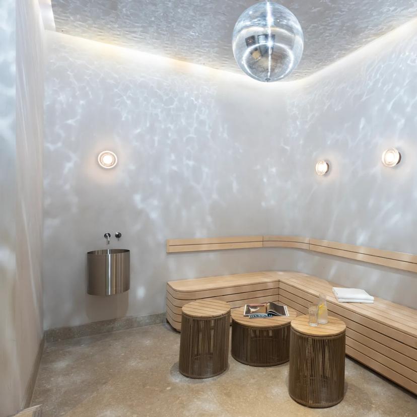 Una moderna sauna con panche in legno, piccoli tavolini rotondi, applique, un asciugamano e un libro. I riflessi di una palla da discoteca creano giochi di luce sulle pareti e sul soffitto.