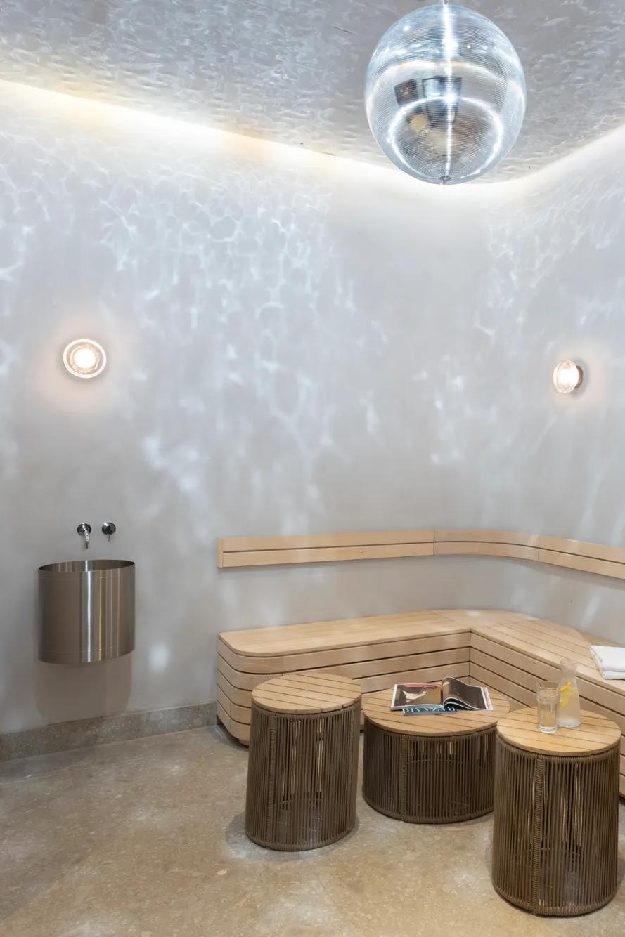 A modern sauna room with wooden benches, small round tables, wall lights, a towel, and a book. Reflections from a disco ball create light patterns on the walls and ceiling.