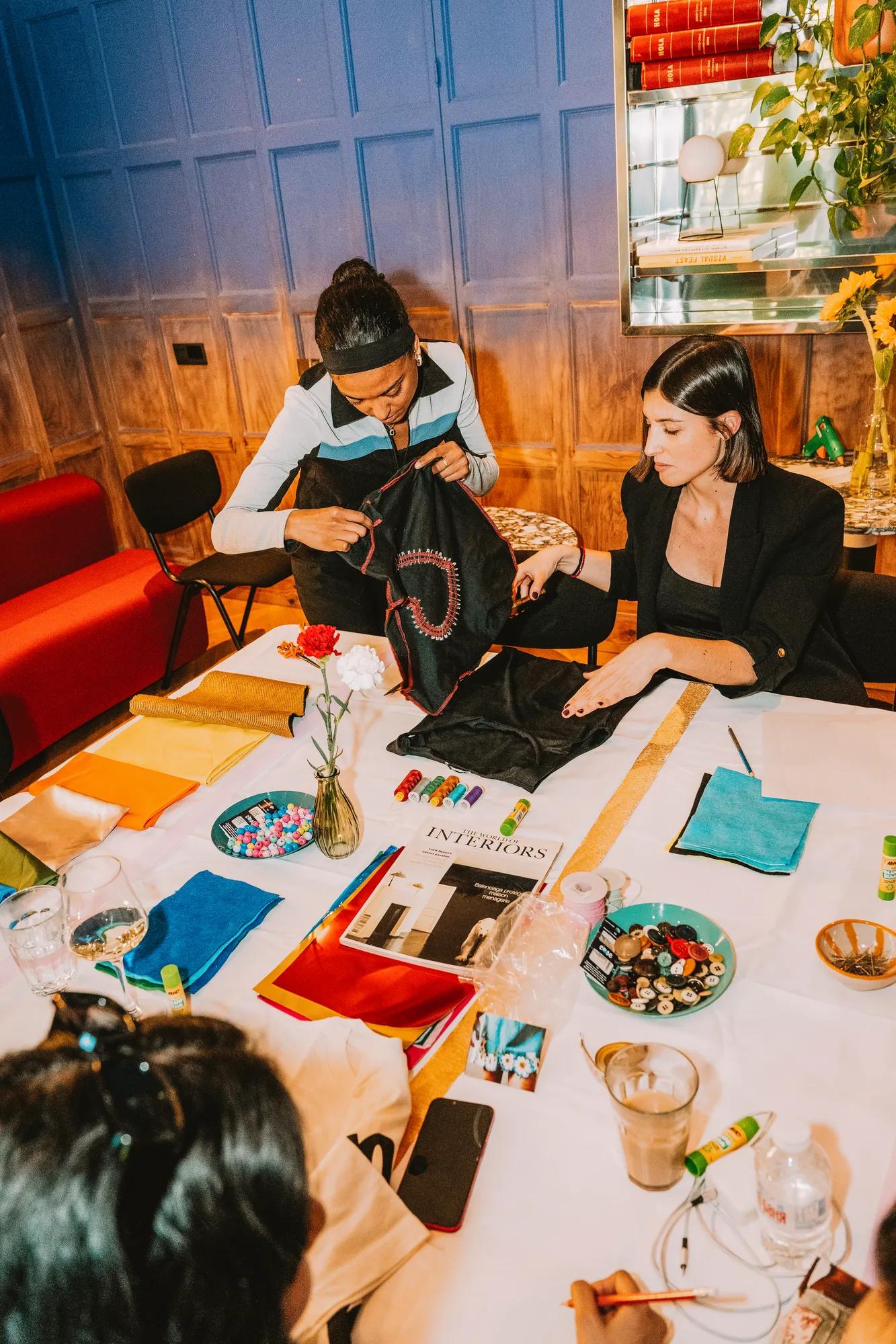Four people sit around a table covered with fabric, beads, and magazines, working on crafts in a cozy, colorful room with wood paneling and a vase of flowers.