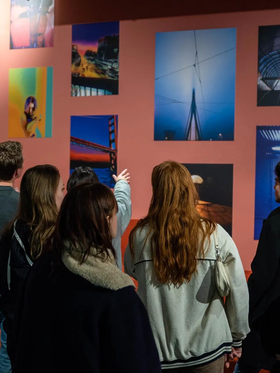 A group of people view colorful photographs displayed on a gallery wall; one person points to an image while others observe, all indoors under warm lighting.