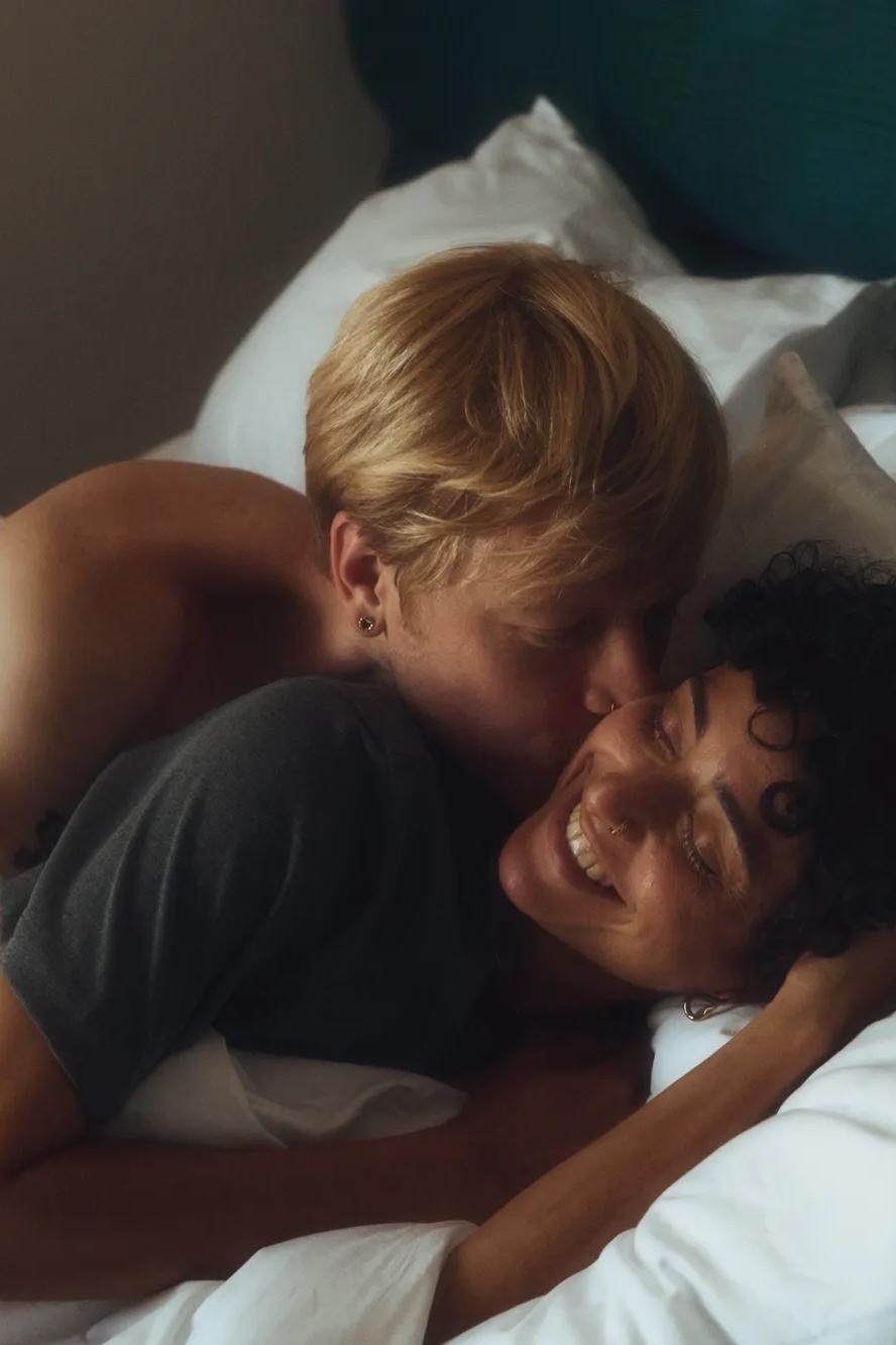 Two people cuddle in bed, smiling and embracing under white blankets. One kisses the other on the cheek as they both appear happy and relaxed. The mood is intimate and affectionate.