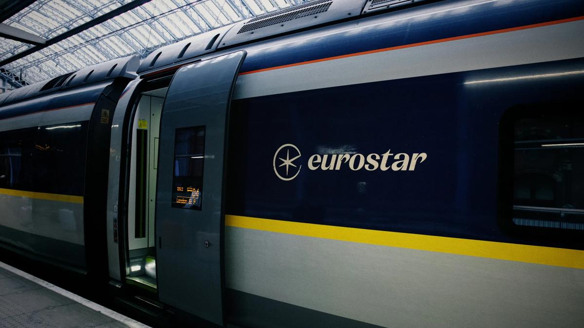 A Eurostar train with an open door is stopped at a platform in a covered station. The train features the Eurostar logo and name on its side, with blue and yellow stripes along the carriage.