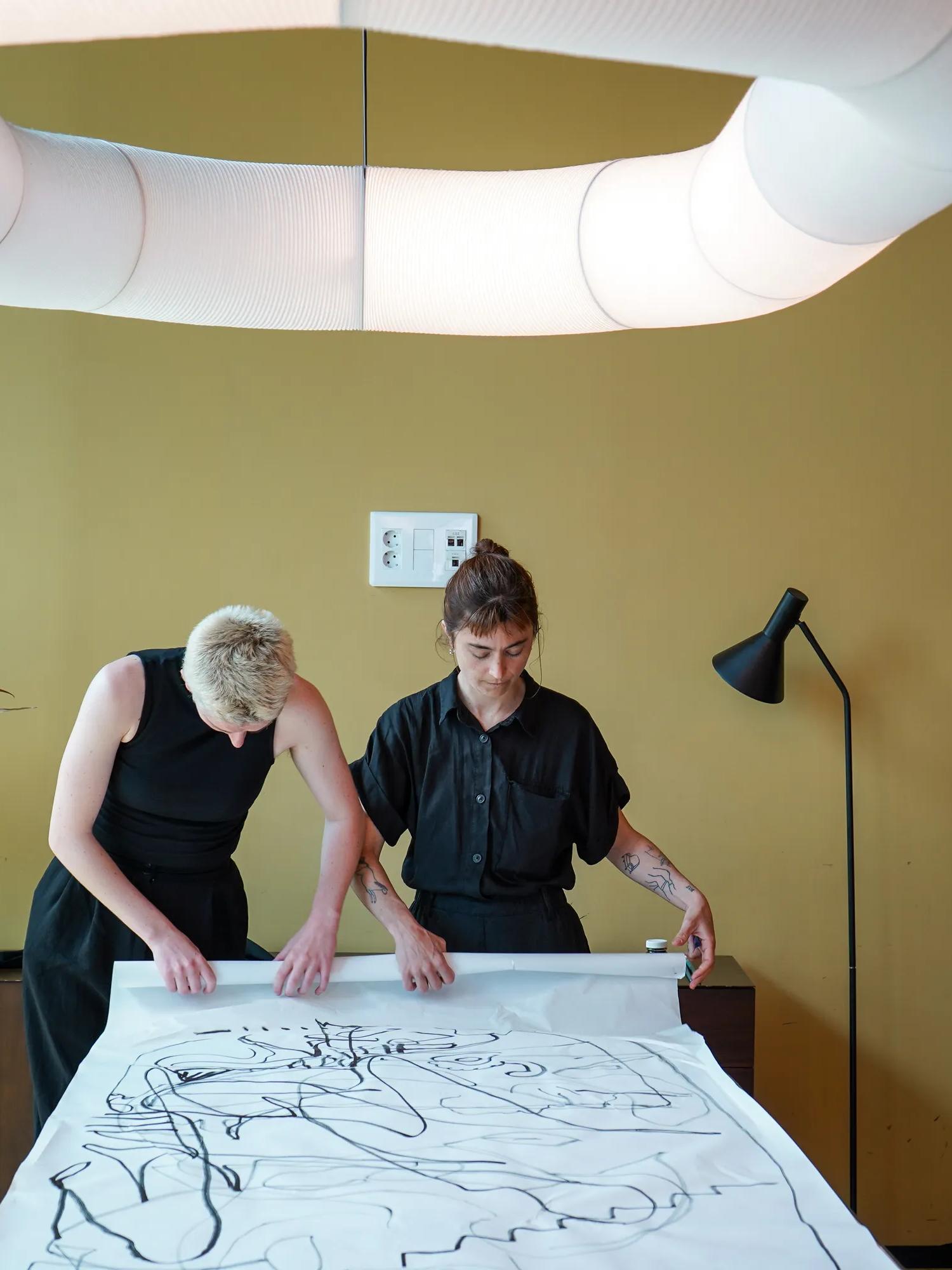 Two people examine a large abstract drawing on a table under modern ceiling lights in a room with a yellow wall, a lamp, and a potted plant.