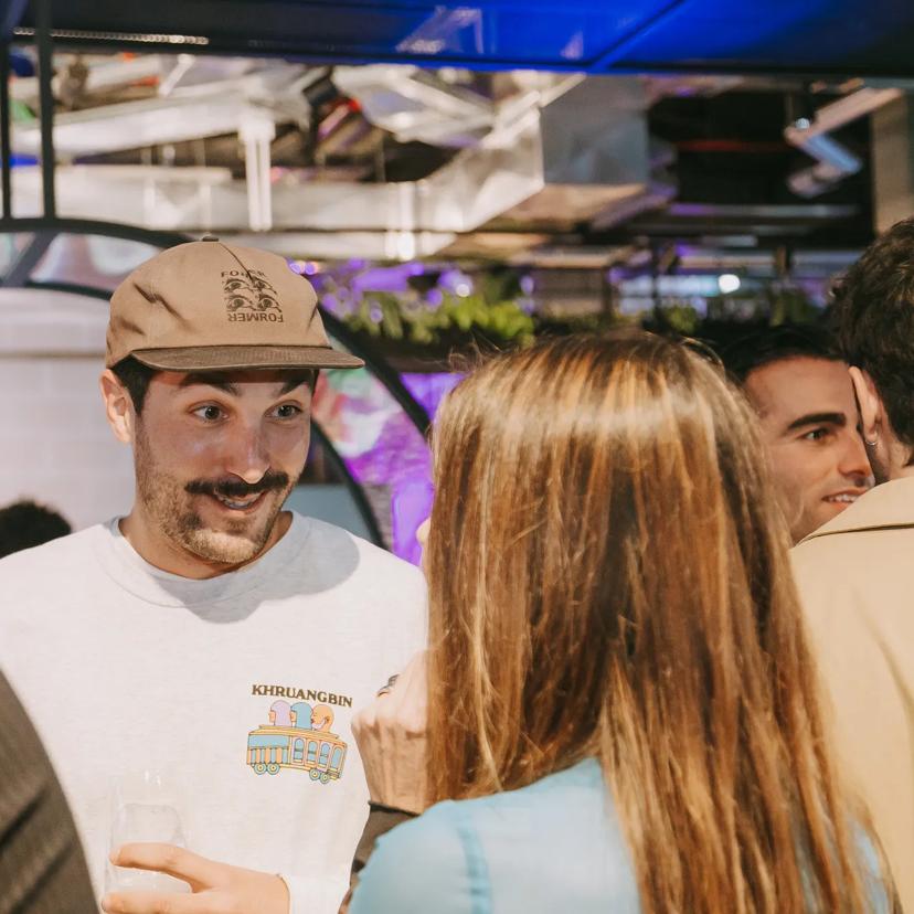 Un hombre, vestido con una sudadera clara y una gorra, sonríe mientras habla con un grupo de personas en un evento social que se celebra en un recinto cerrado, con una bebida en la mano.