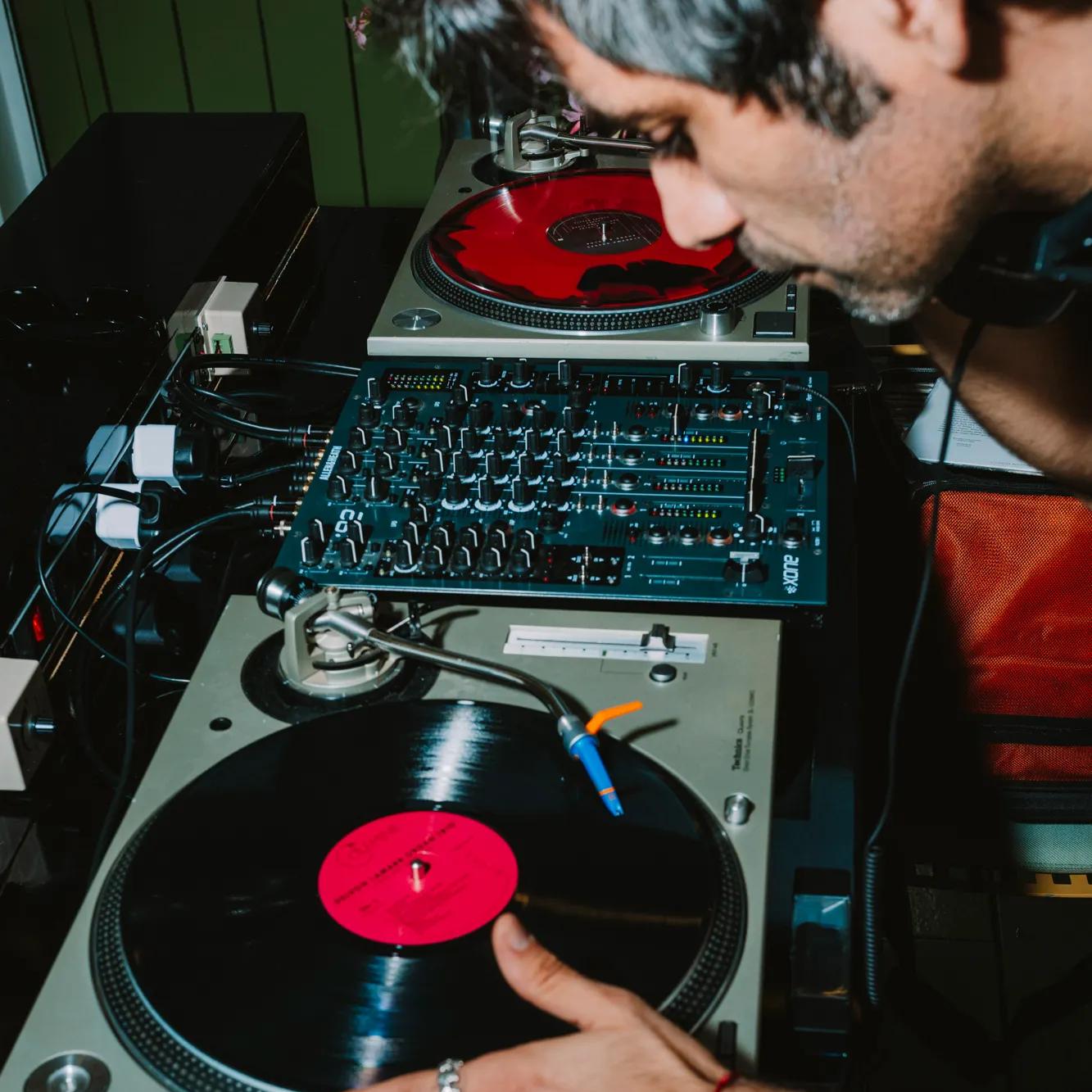 A person uses a turntable, adjusting a vinyl record with a red label, next to a mixer and another turntable, in a dimly lit setting.