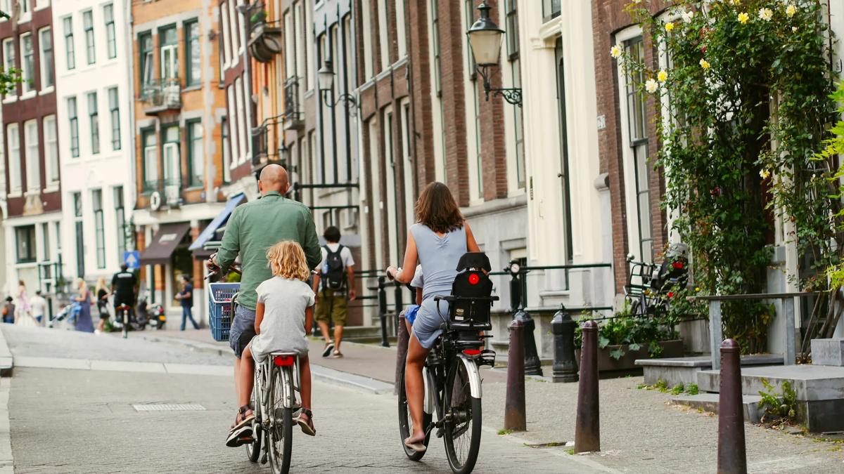 Three people ride bicycles on a city street lined with tall, narrow buildings. Two children and an adult are seen from behind, cycling together on a sunny day.