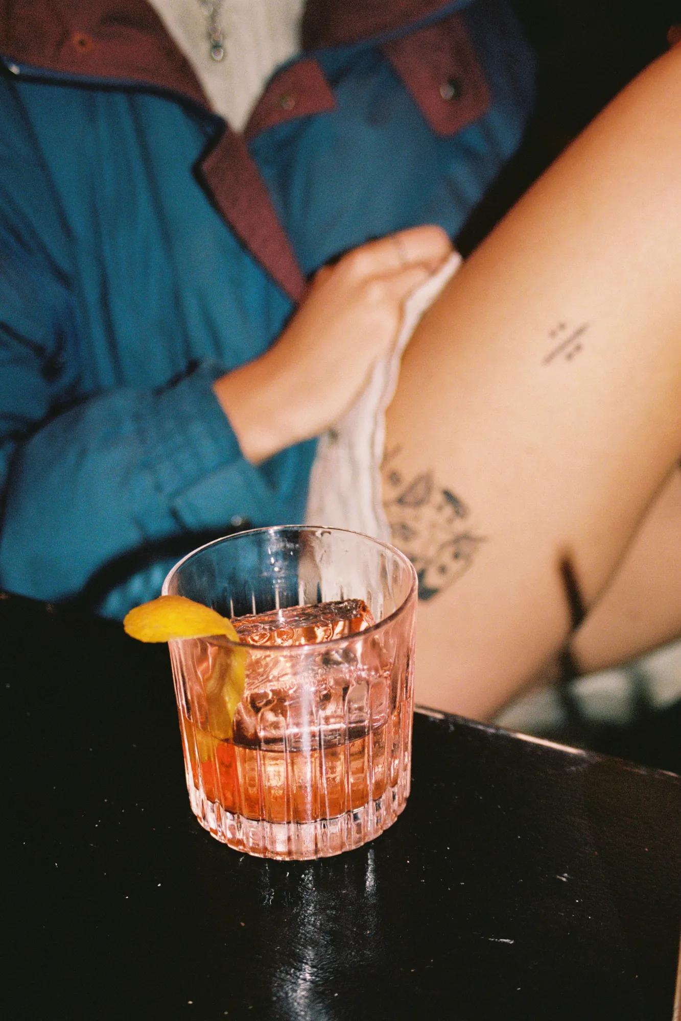 A glass of cocktail with an orange peel garnish sits on a dark table. In the background, a person in a blue jacket lifts their skirt, revealing a tattoo on their thigh.