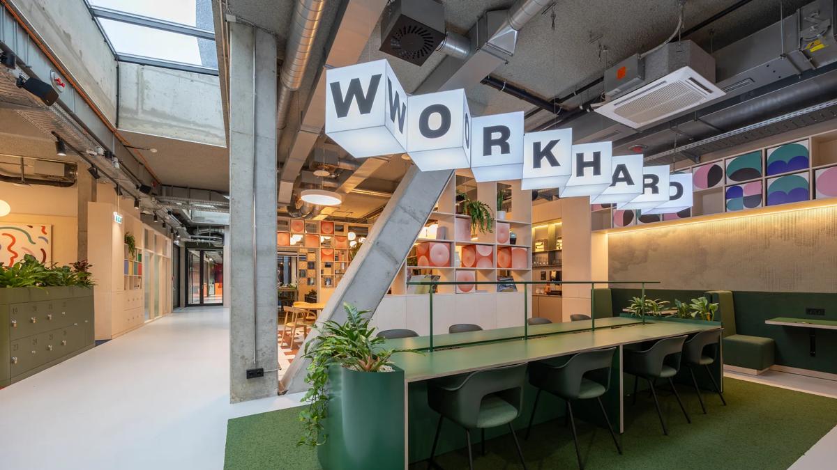 Modern open office space with green desks, plants, cube lights spelling WORK HARD, exposed ceiling pipes, and colorful wall art, creating a stylish and vibrant work environment.