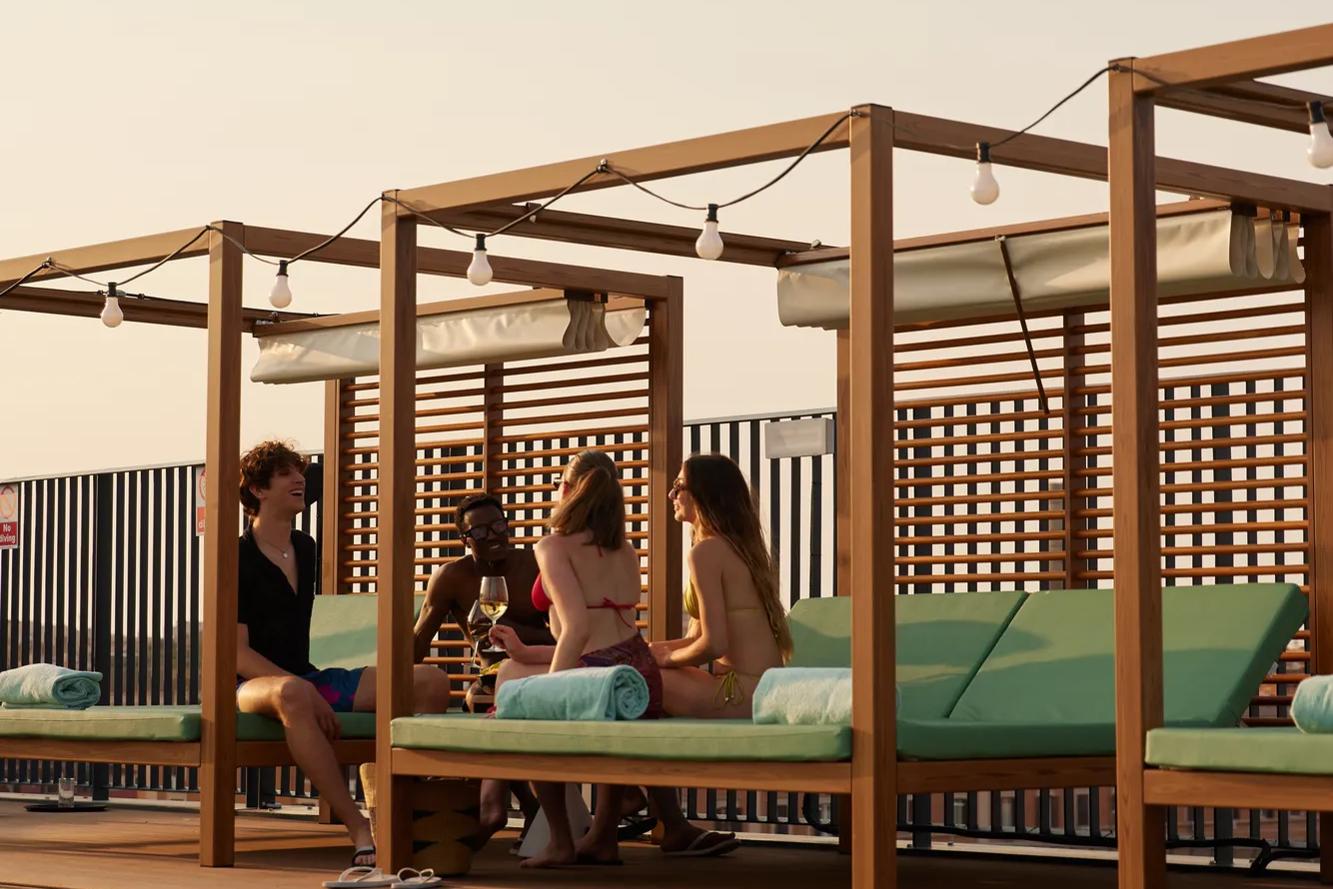 Quatro pessoas estão sentadas conversando em espreguiçadeiras sob um guarda-sol à beira de uma piscina, com a água azul em primeiro plano e uma cerca de madeira ao fundo.