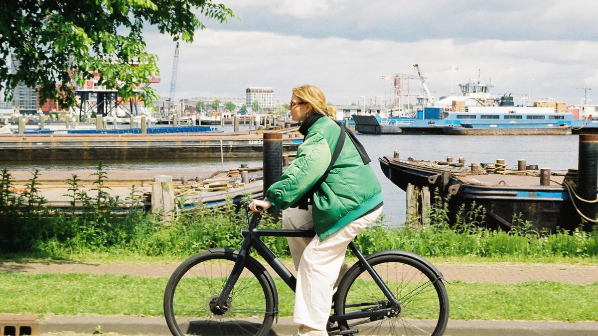 A person in a green jacket rides a black bicycle along a waterfront path, with boats and industrial structures visible across the water in the background.