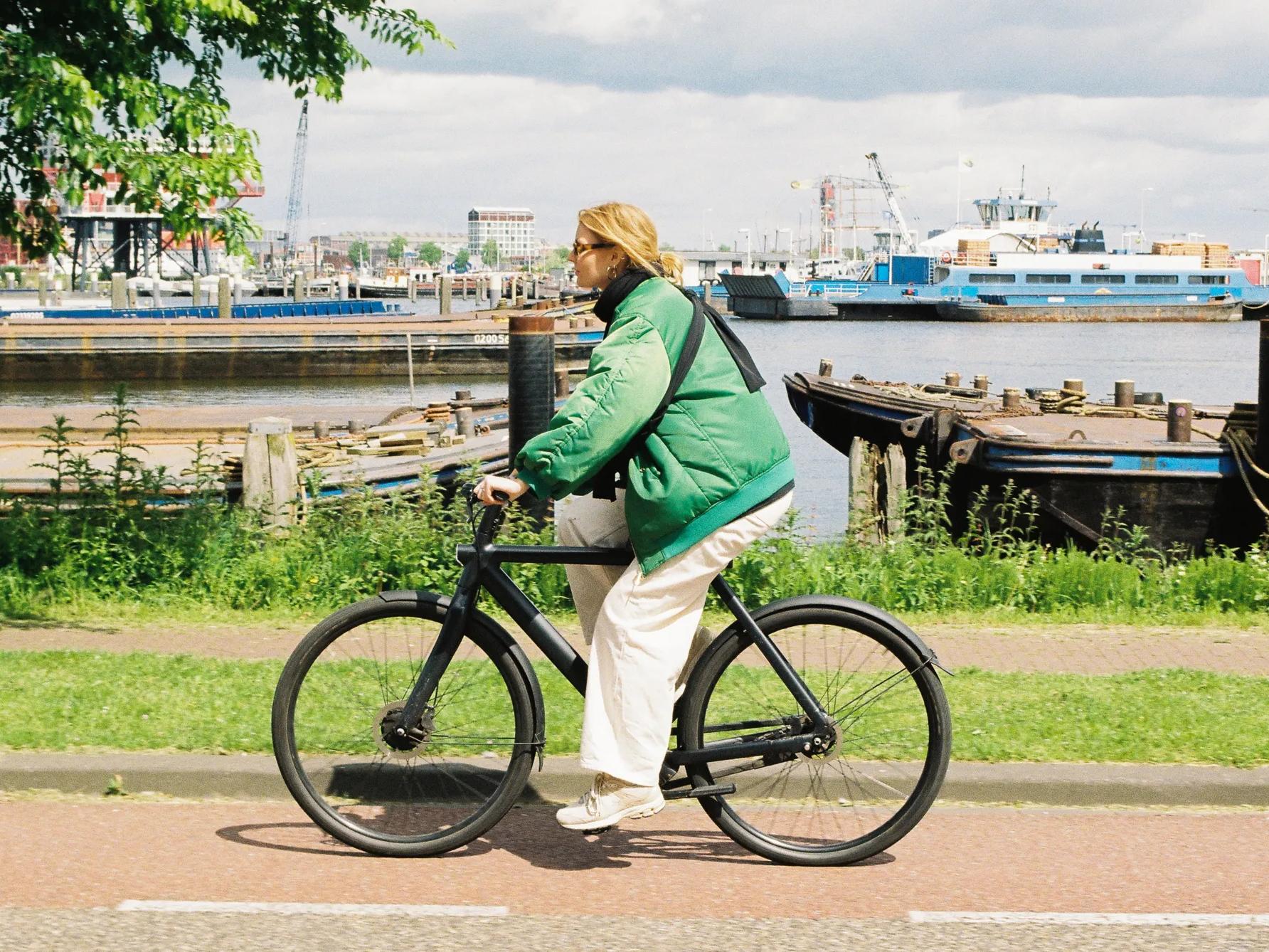 Una persona con una chaqueta verde va en bicicleta negra por un camino junto al río, con barcos y estructuras industriales visibles al otro lado del agua, al fondo.