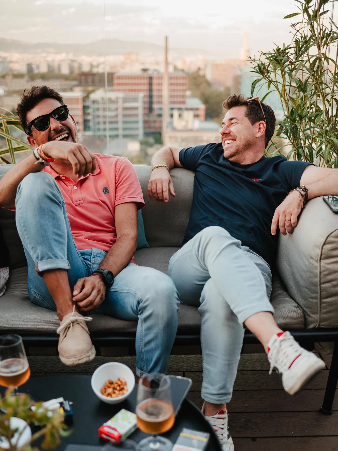Two men sit on an outdoor couch, laughing and relaxing with drinks and snacks. A cityscape is visible in the background, and plants surround them.