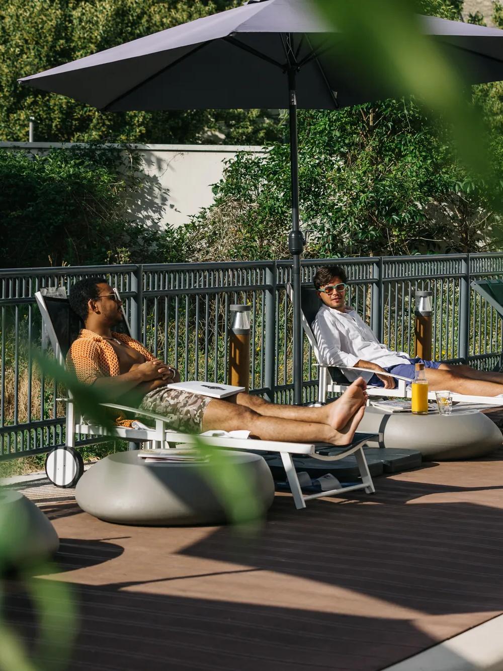 Two people relax on lounge chairs by a pool, shaded by an umbrella. They each have drinks nearby and are surrounded by greenery and empty chairs. Leaves partially frame the foreground.