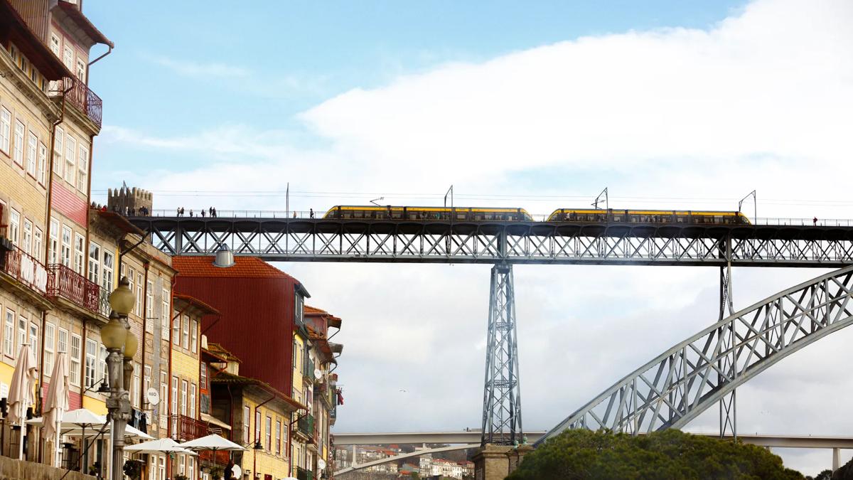 A yellow tram crosses a high metal bridge above colorful buildings and outdoor cafes in a riverside city, under a partly cloudy sky.