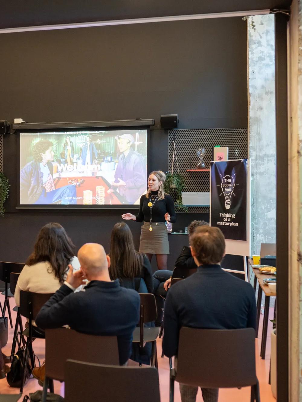 A woman gives a presentation to a seated audience in a modern classroom. A skeleton model stands nearby, and a screen displays the word Medical. The room has exposed concrete and colorful decor.