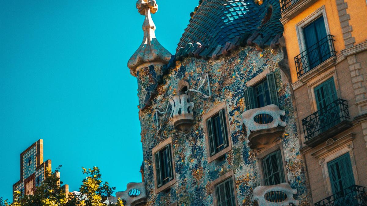 Colorful, mosaic-covered building with wavy lines, curved balconies, and a unique tiled roof, set against a clear blue sky. Neighboring buildings have traditional balconies and straight lines.