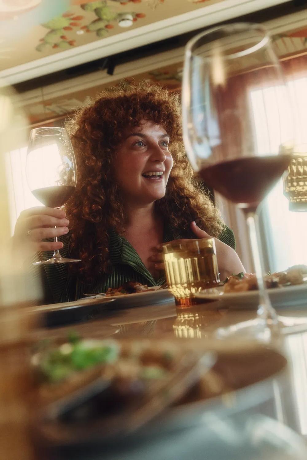 Two women sit at a table, smiling and enjoying drinks and food together in a warmly lit restaurant. One holds a glass of red wine while the other has a drink in a faceted glass.