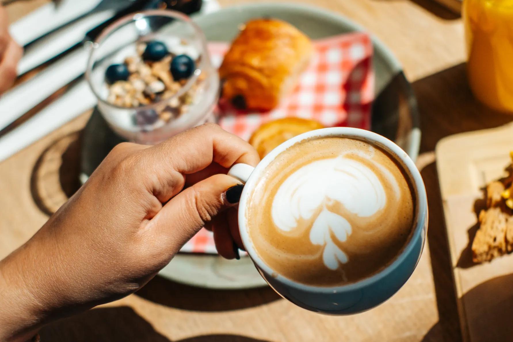 A hand holds a ceramic mug of latte with heart-shaped foam art above a table set with a croissant, a glass of yogurt with blueberries, and glasses of orange juice.