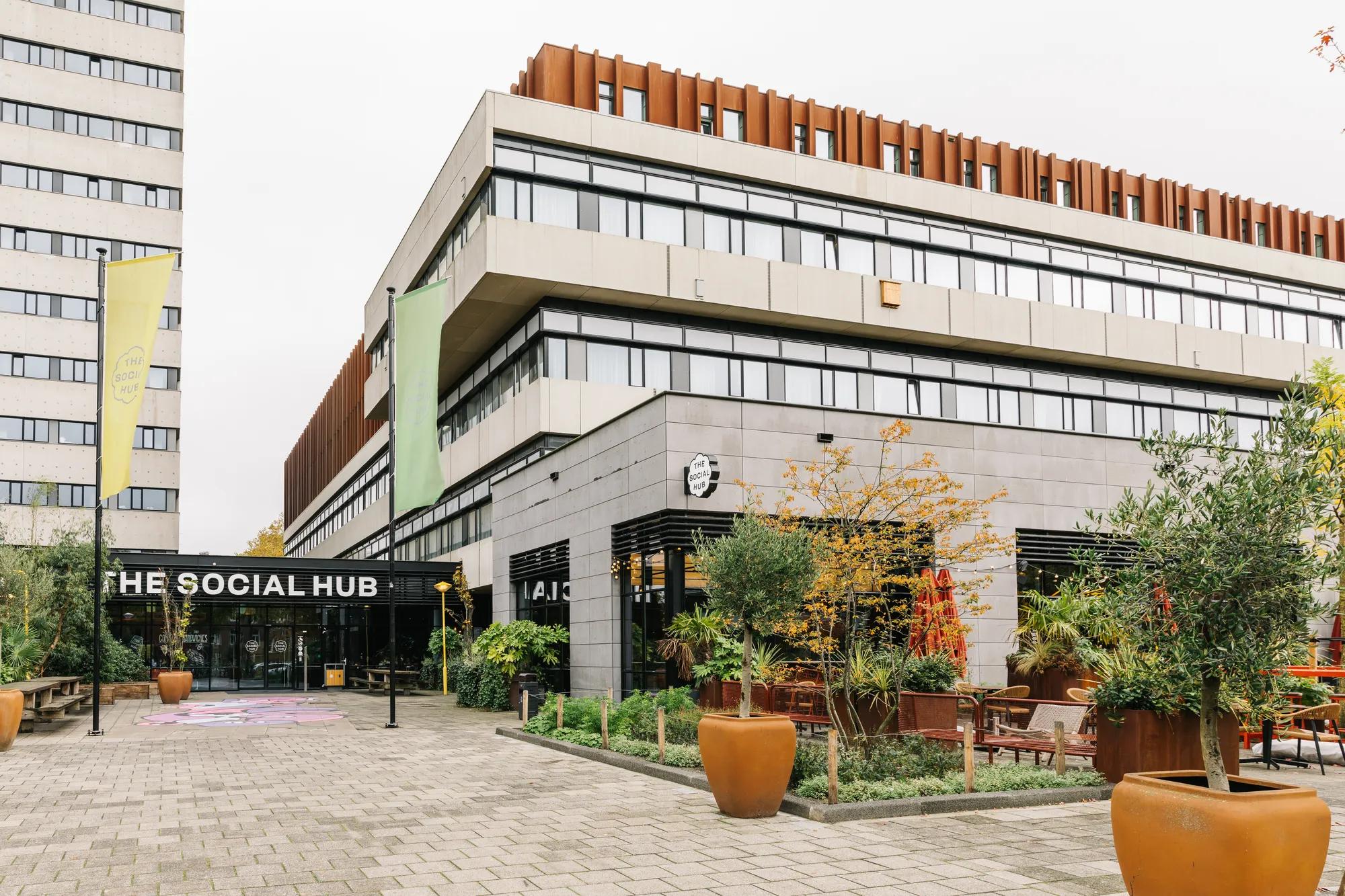 Modern building with large windows, labeled The Social Hub at the entrance. Outdoor seating, potted plants, and greenery decorate the paved area in front. Tall building visible to the left in the background.