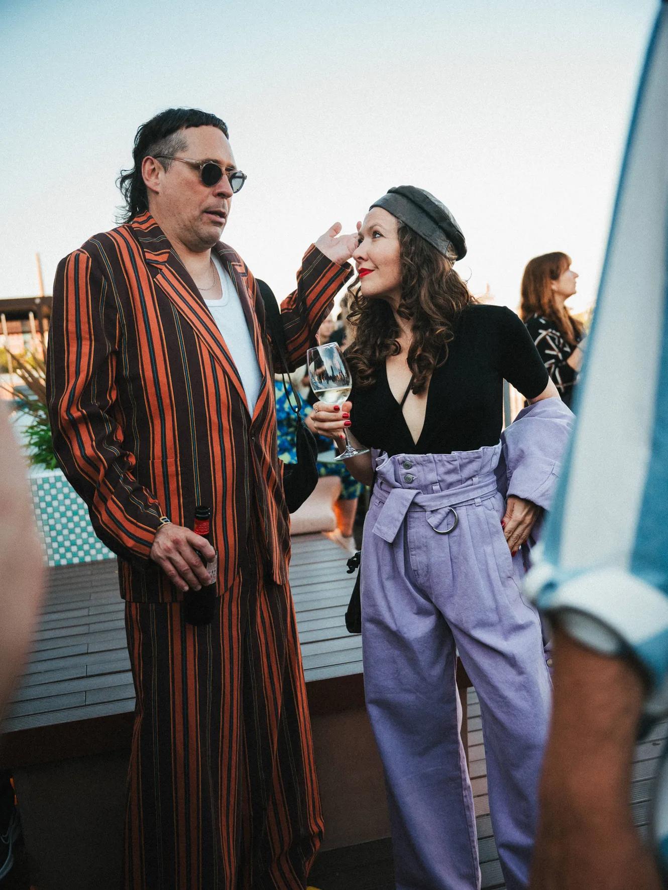 A man in a striped suit and sunglasses talks to a woman in a black top, purple high-waisted pants, and a gray beret. Both hold drinks at an outdoor gathering.