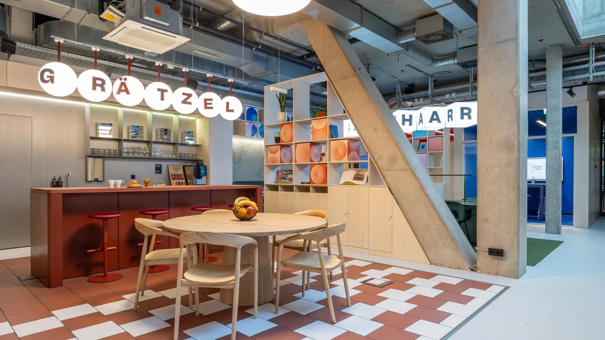 Modern office kitchen and dining area with red and white tiled floor, wooden table, red chairs, open shelves, and bold round signage reading GRÄTZEL and HAARR under exposed ceilings and ductwork.