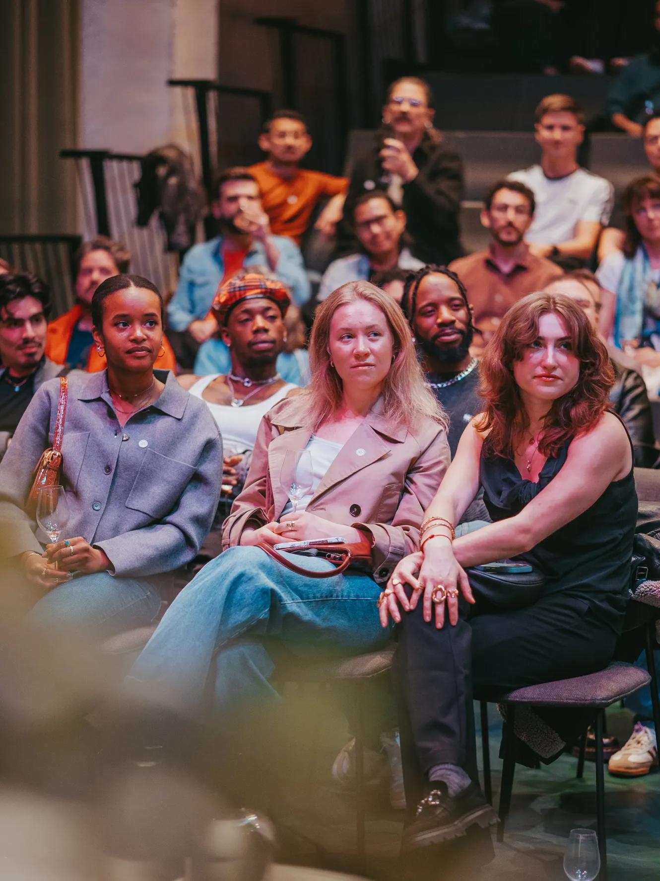 A diverse group of people sits closely together, attentively watching an event or presentation in an indoor setting. The atmosphere appears engaged and focused.
