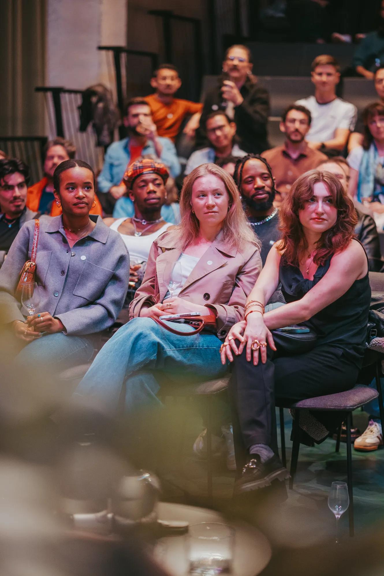 A diverse group of people sits closely together, attentively watching an event or presentation in an indoor setting. The atmosphere appears engaged and focused.