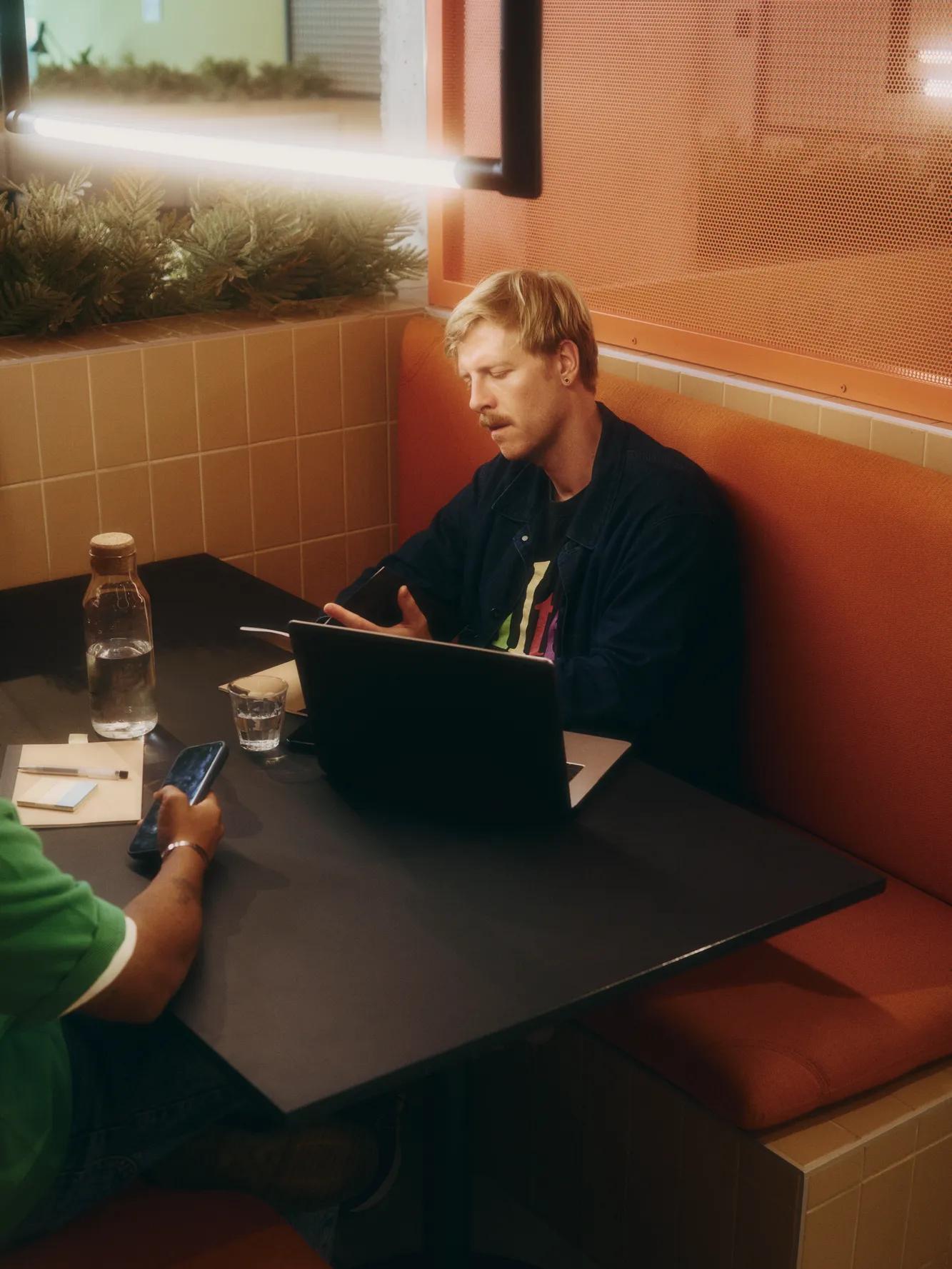 Two people sit in an orange booth at a café. One works on a laptop, talking, while the other holds a phone. Theres a glass water bottle and glasses on the table.