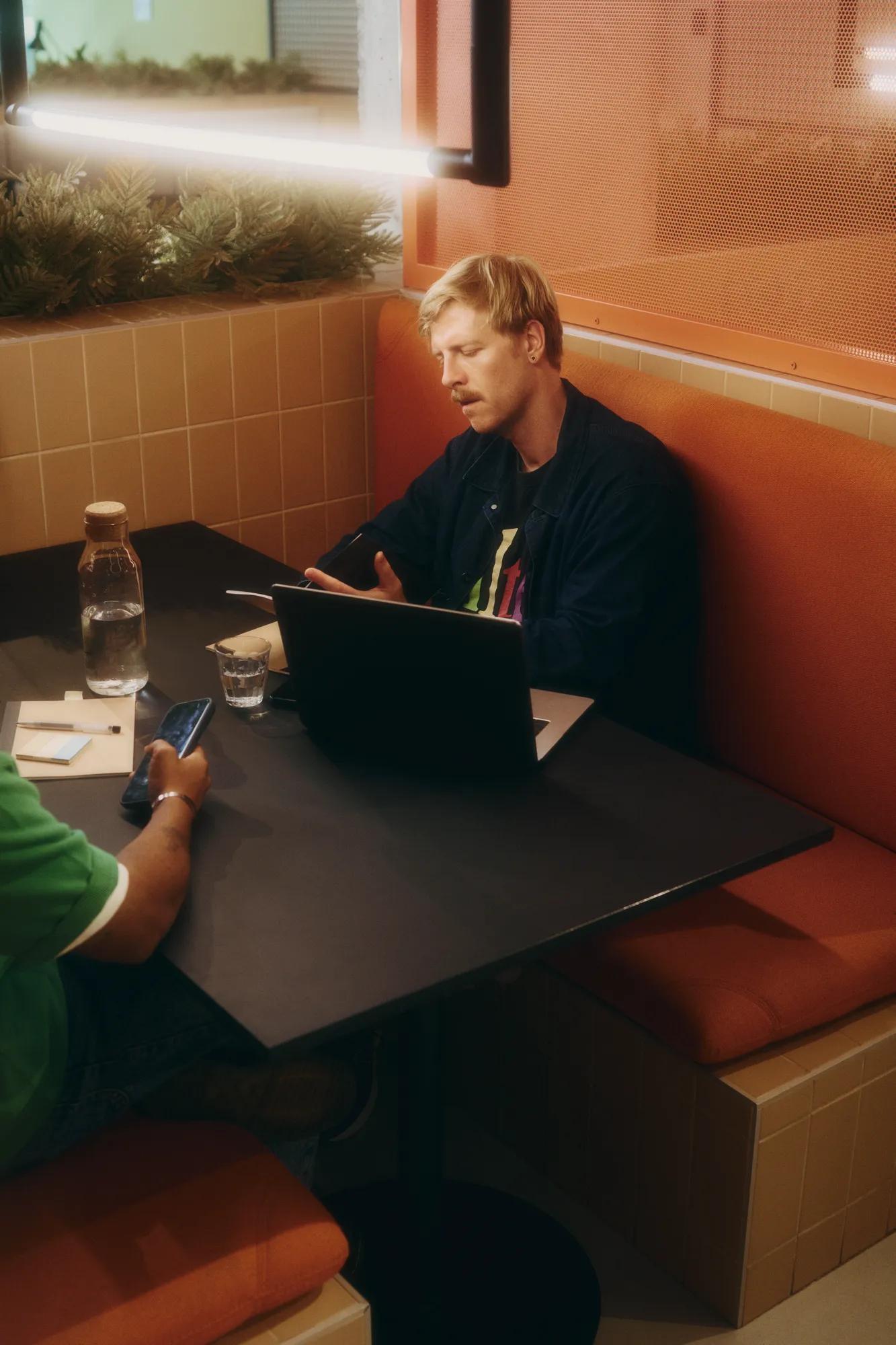 Two people sit in an orange booth at a café. One works on a laptop, talking, while the other holds a phone. Theres a glass water bottle and glasses on the table.