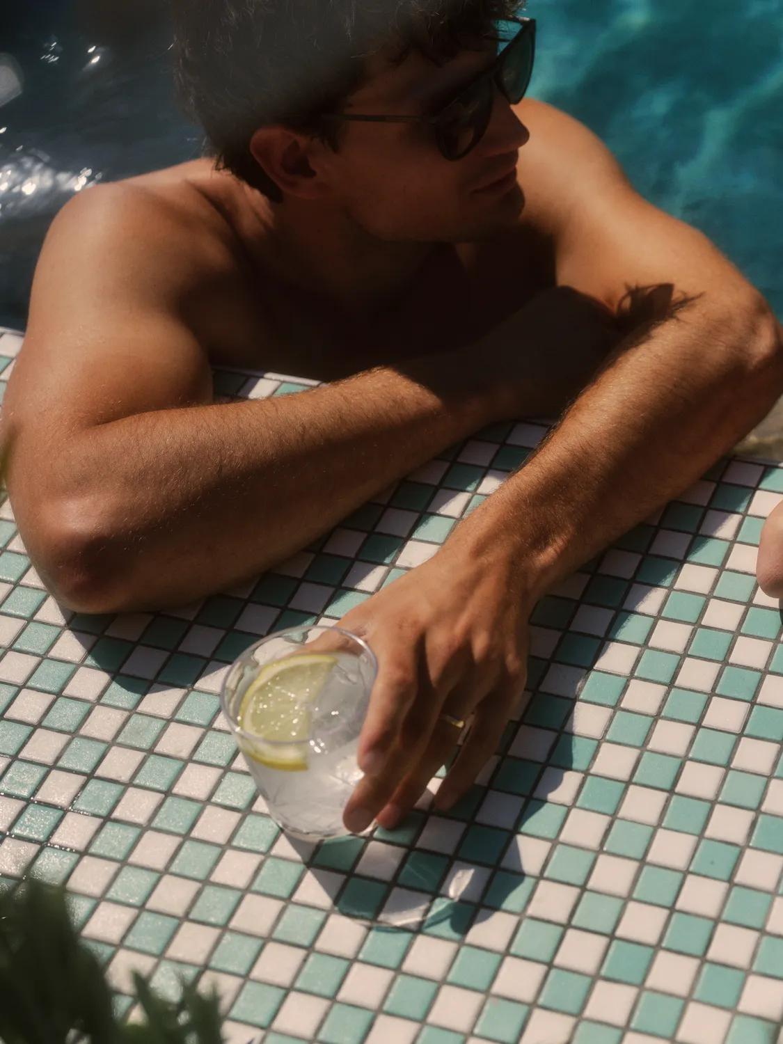 Two people relax at the edge of a tiled swimming pool. One holds a drink with lemon slices. They both wear sunglasses, and one person wears a sunhat. Sunlight creates reflections on the water.