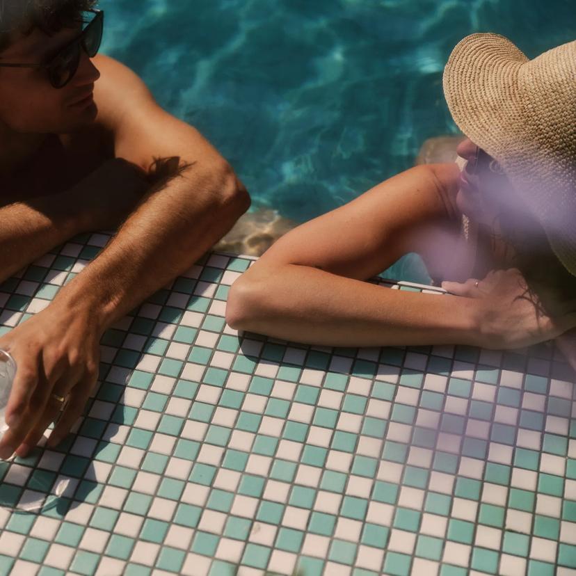Two people relax at the edge of a tiled swimming pool. One holds a drink with lemon slices. They both wear sunglasses, and one person wears a sunhat. Sunlight creates reflections on the water.