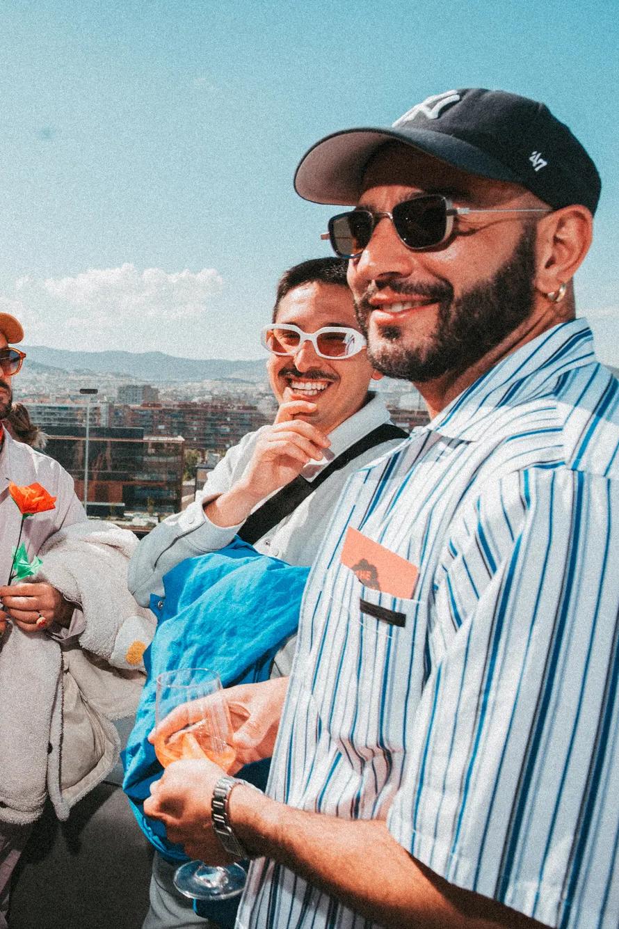 Five people stand together on a sunny rooftop, all wearing sunglasses and trendy outfits, smiling and enjoying drinks with a cityscape and blue sky in the background.