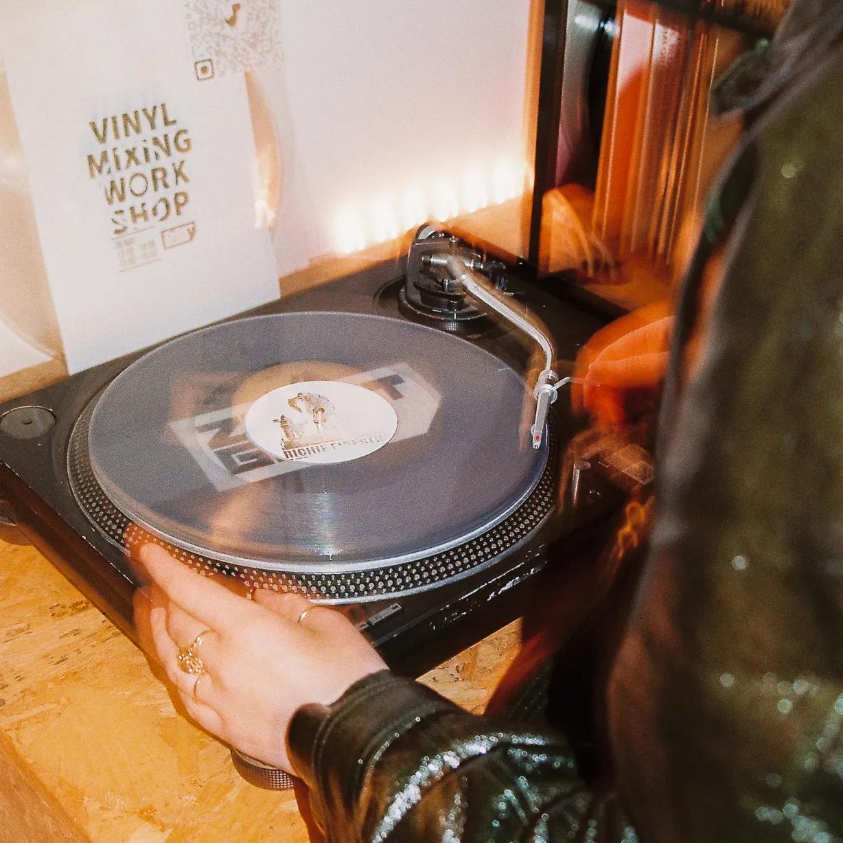 A person in a dark jacket adjusts a vinyl record on a turntable. A sign reading Vinyl Mixing Workshop is visible nearby, along with shelves holding records. The setting appears to be a cozy, creative workspace.