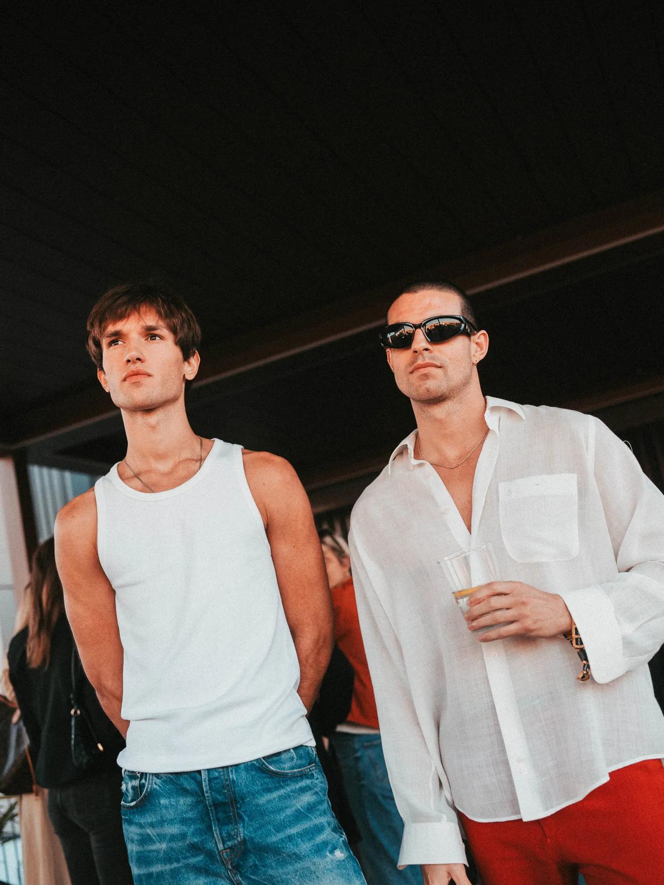 Two men stand indoors; one wears a white tank top and jeans, the other wears sunglasses, a sheer white shirt, and red pants, holding a cup. People are blurred in the background.