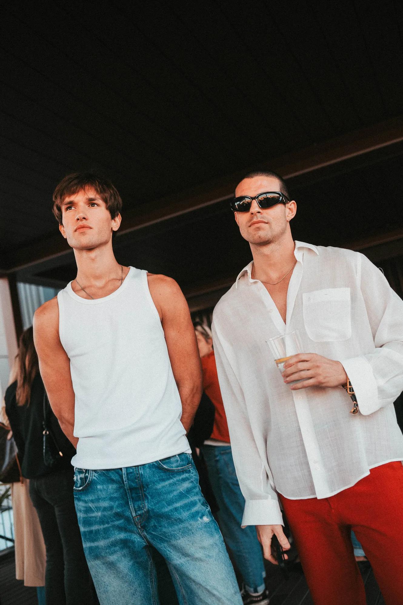 Two men stand indoors; one wears a white tank top and jeans, the other wears sunglasses, a sheer white shirt, and red pants, holding a cup. People are blurred in the background.