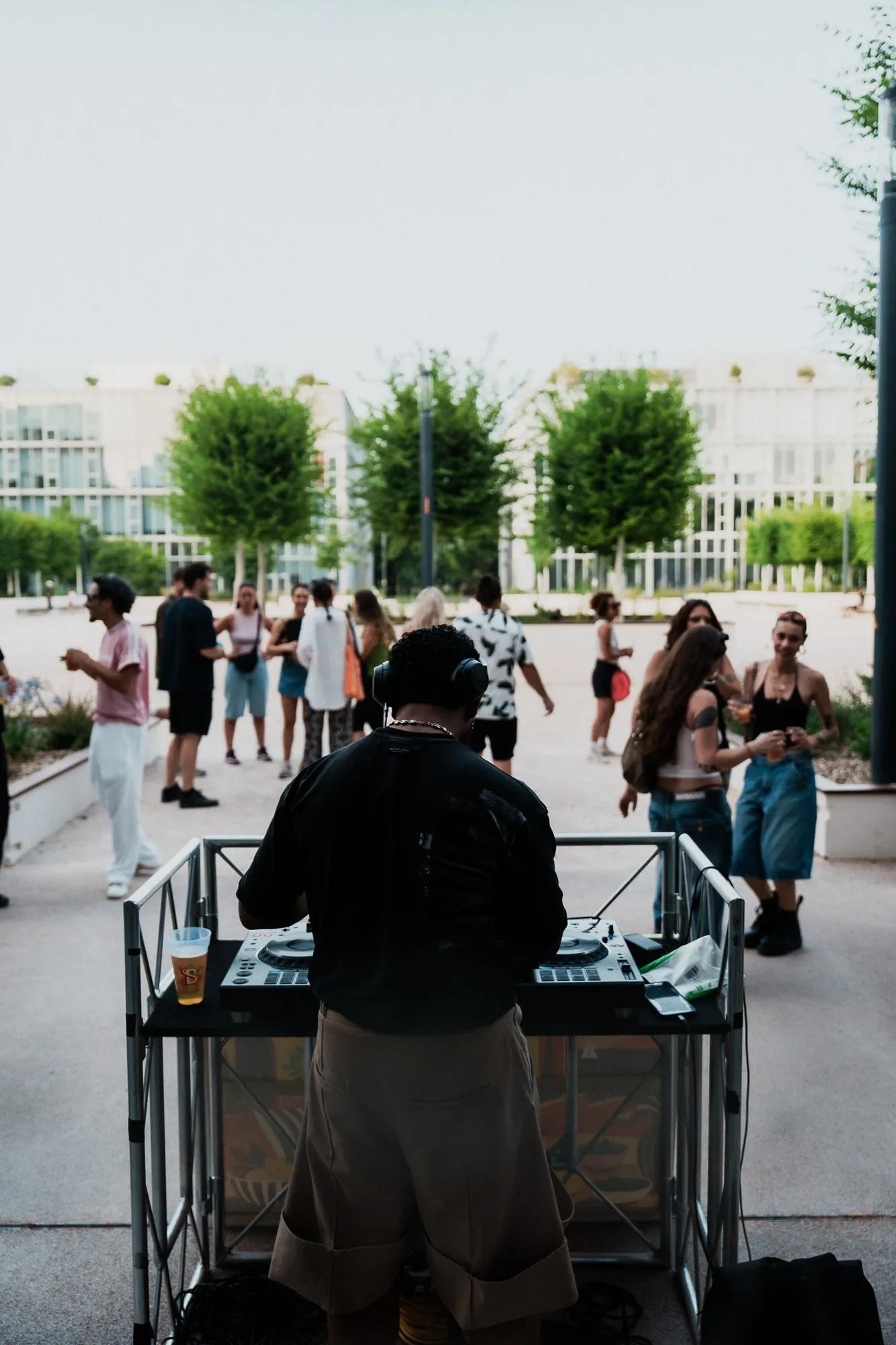 Ein DJ legt im Freien Musik auf, während im Hintergrund Menschen tanzen und sich unterhalten. Im Hintergrund sind Bäume und moderne Gebäude zu sehen, und auf dem Tisch des DJs steht ein Getränk.