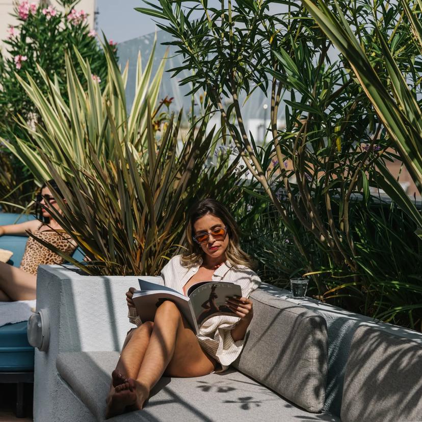 Three people relax outdoors among lush plants. Two lounge on blue sunbeds; one wears a swimsuit and sunbathes. The other sits on a couch, wears sunglasses, and reads a magazine. Sunlight filters through the greenery.
