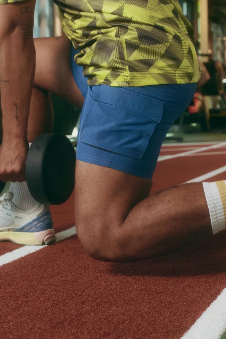 Person in blue shorts and colorful socks kneels on a track indoors, holding a dumbbell in one hand, preparing for an exercise in a gym with equipment visible in the background.