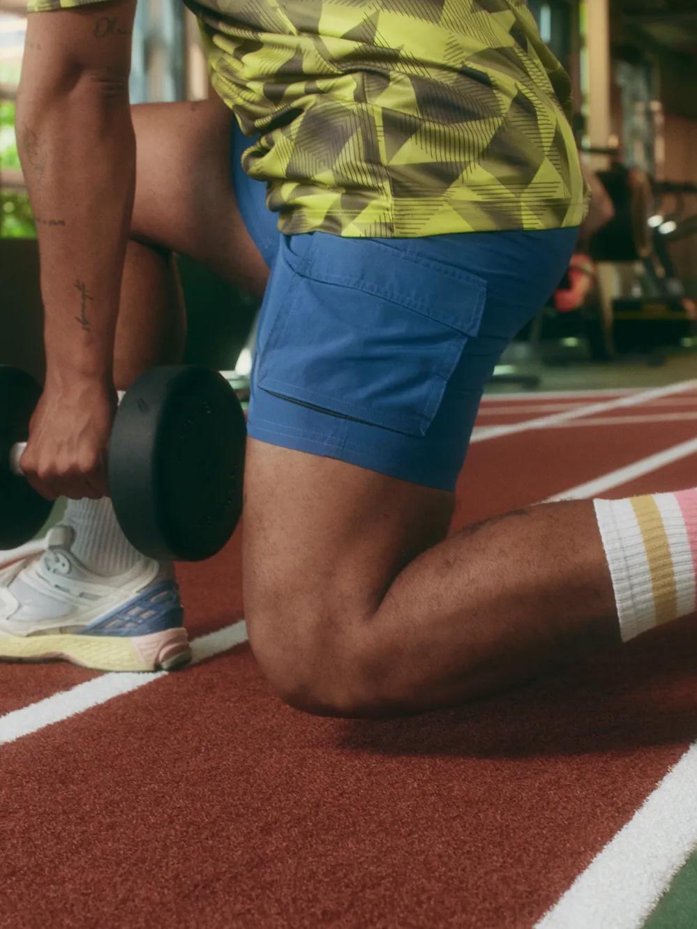 Person in blue shorts and colorful socks kneels on a track indoors, holding a dumbbell in one hand, preparing for an exercise in a gym with equipment visible in the background.
