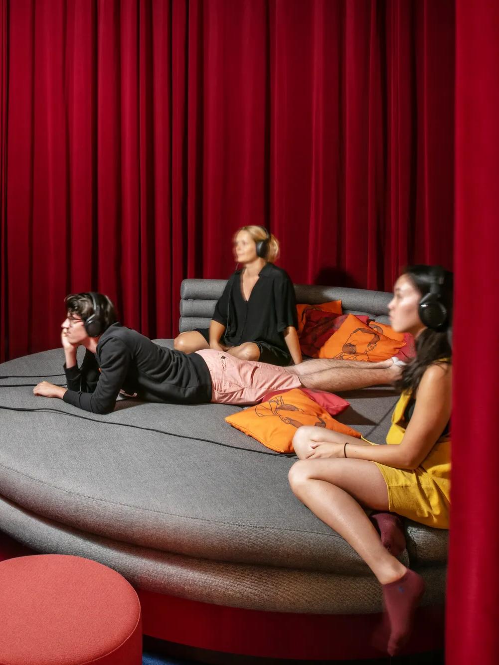 Three people with headphones sit and recline on a round, gray couch with orange pillows, watching a screen in a room with red curtains and a dark blue carpet.