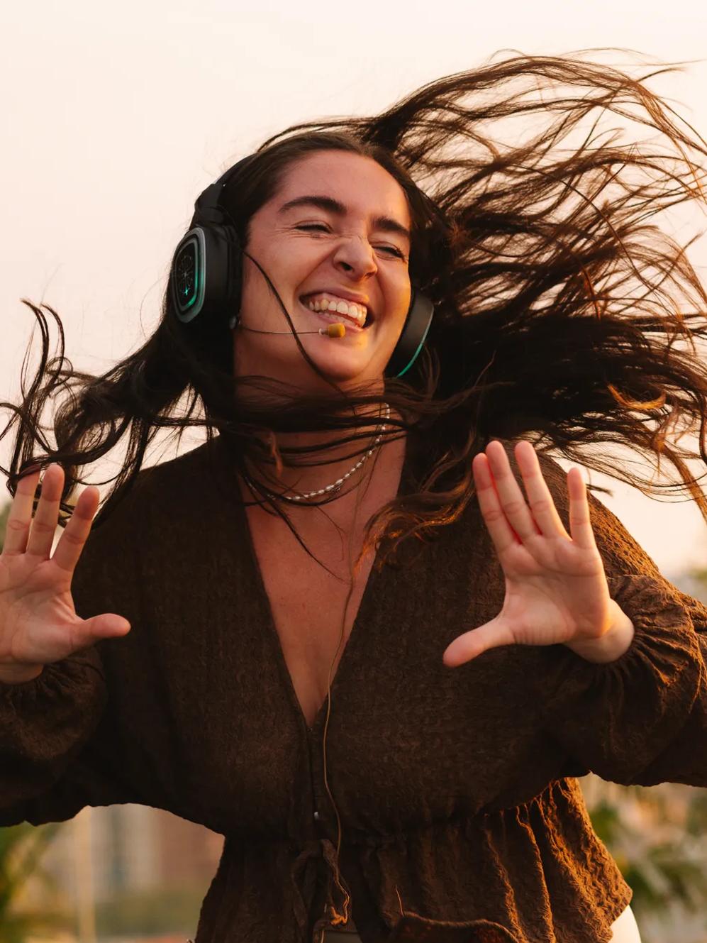 A woman with long, flowing hair dances joyfully outdoors while wearing headphones, smiling with her eyes closed and hands raised, enjoying music in the sunlight.