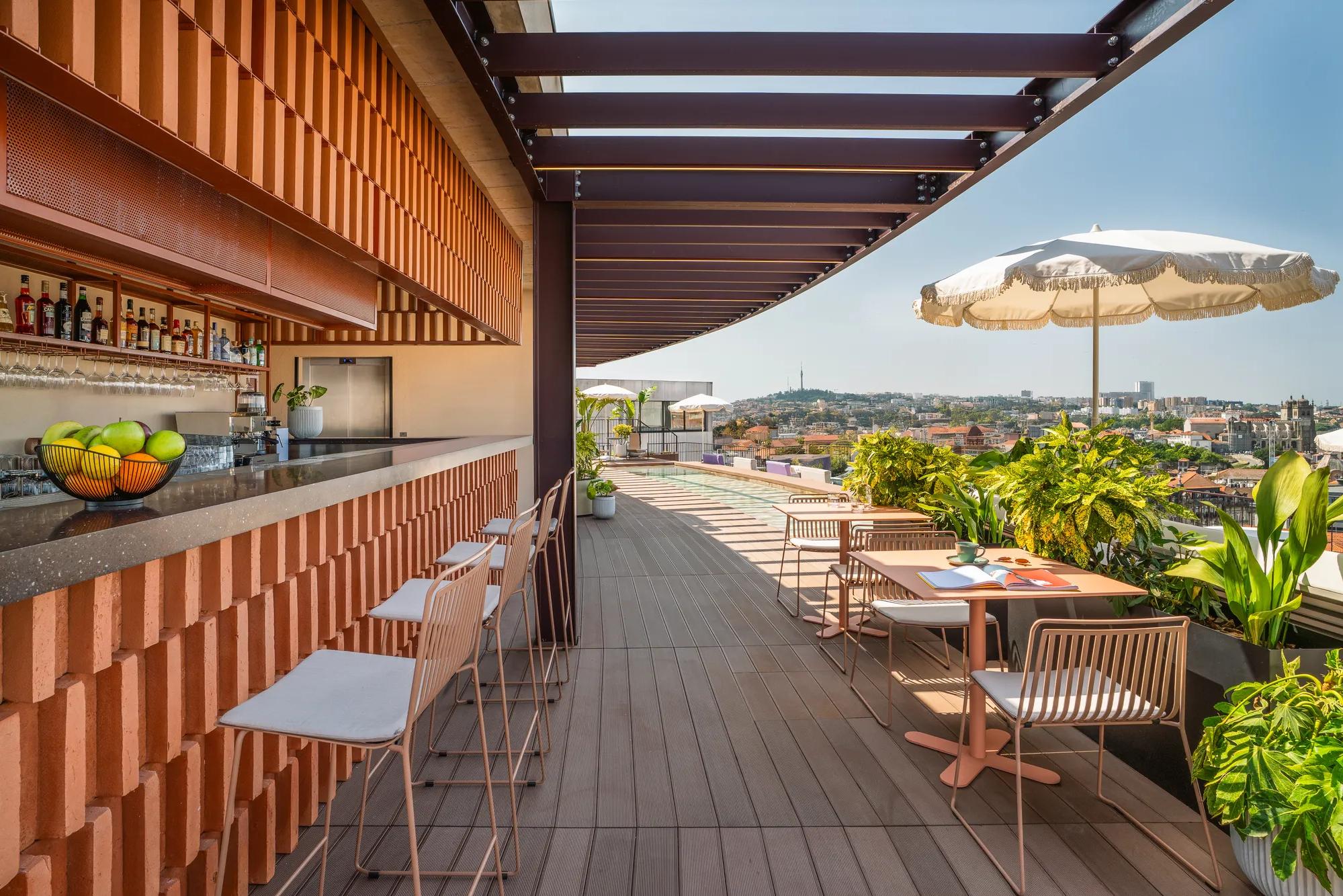 A rooftop bar with a modern design, high chairs at the counter, tables with umbrellas, potted plants, and a city view under a clear blue sky.