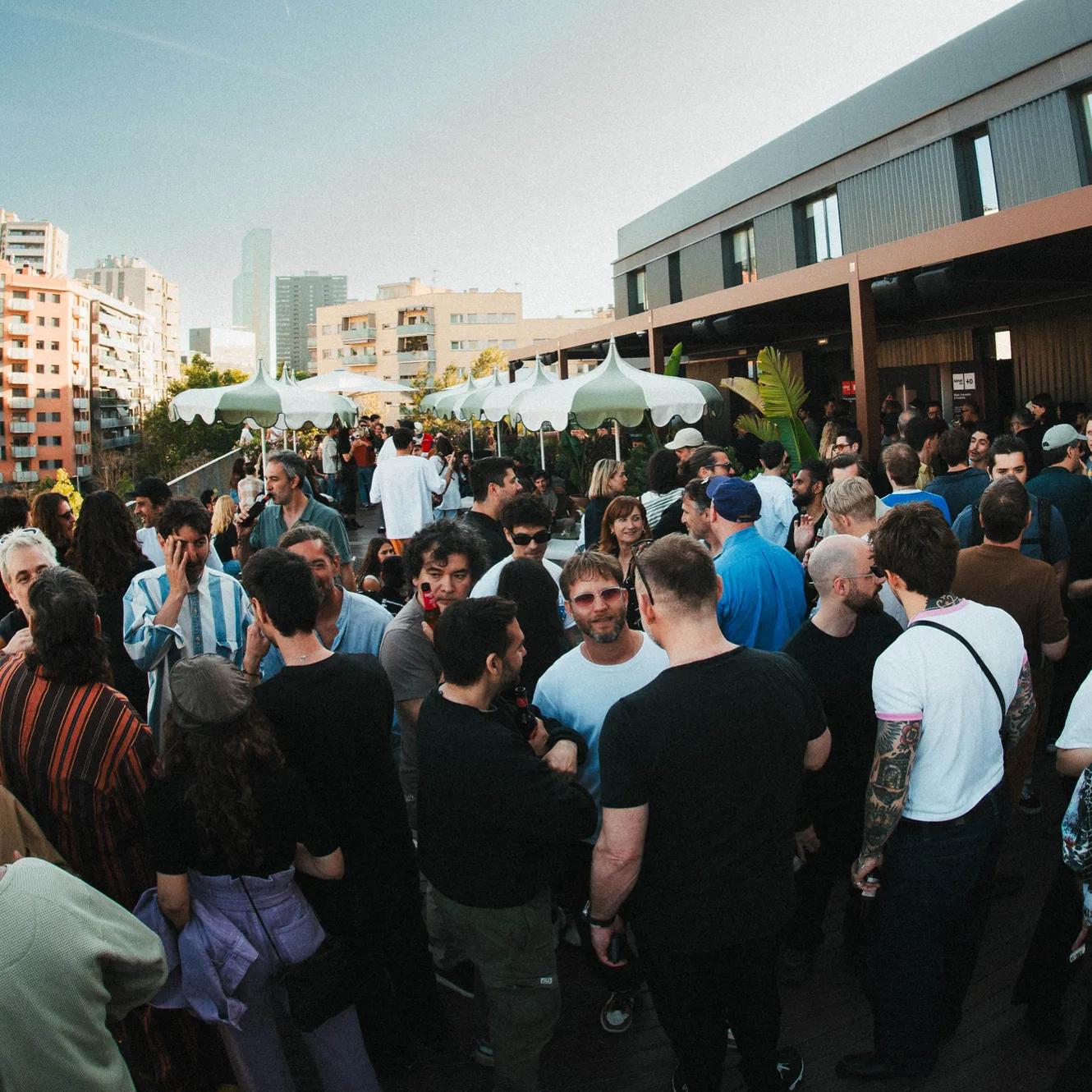 A large group of people socialize at an outdoor rooftop gathering, surrounded by buildings, under a clear blue sky.