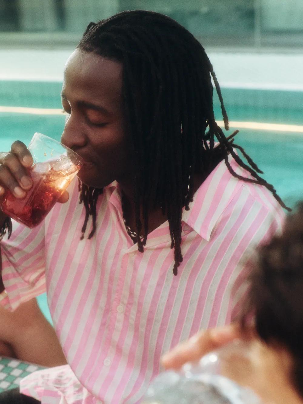 A person in a pink striped shirt drinks from a cup by a pool, surrounded by friends who are also holding drinks. The scene is relaxed and summery.