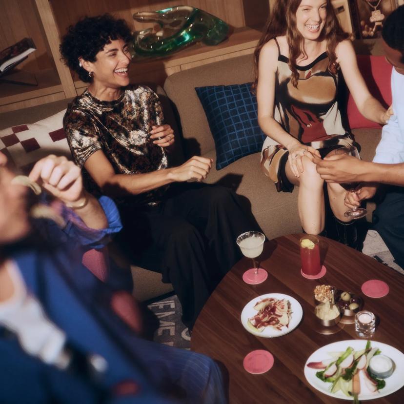 Quatro pessoas estão sentadas juntas em sofás, sorrindo e rindo, ao redor de uma mesa de madeira com coquetéis e pratos de aperitivos, em um ambiente aconchegante.