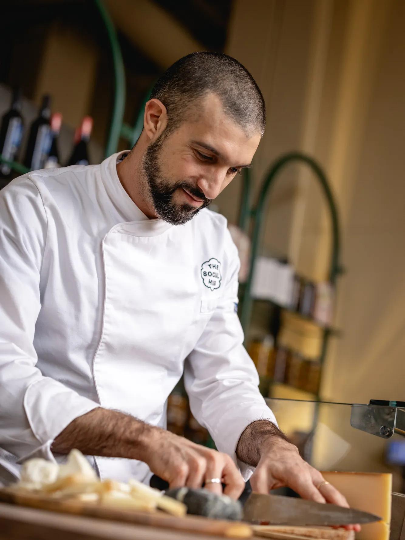 A chef in a white uniform slices food on a wooden counter, smiling and focused, in a warmly lit kitchen with bottles and jars on shelves in the background.